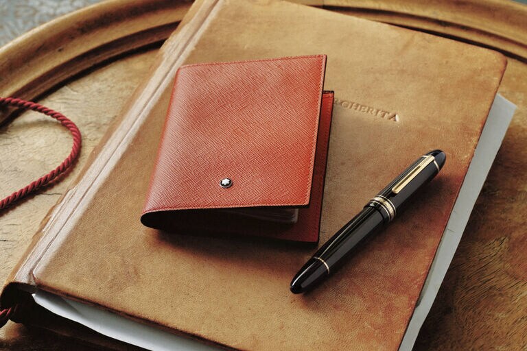 Montblanc Meisterst&uuml;ck fountain pen in black precious resin with gold details, alongside a red leather card holder with snowcap emblem, on a vintage leather notebook.