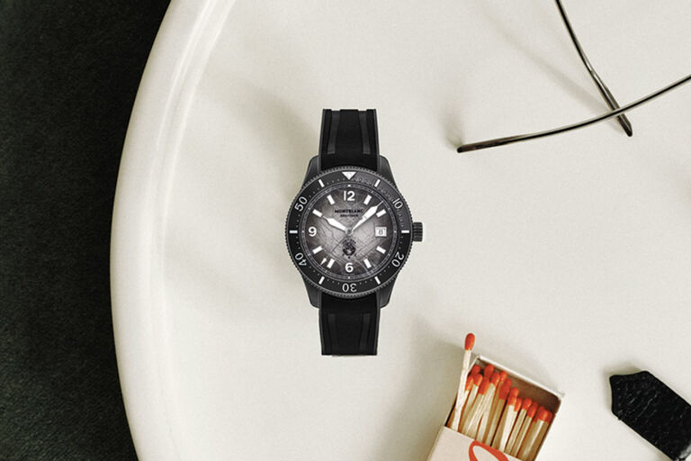Montblanc black dive watch with textured grey dial, luminous markers, and black rubber strap, flat lay on a light surface with eyeglasses.