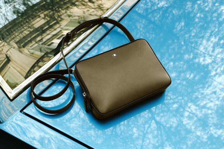 Montblanc leather clutch in dark grey suede with integrated handle, styled on a bright orange towel in a sunlit outdoor setting.